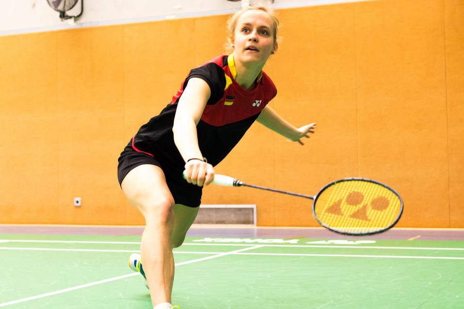 Spielerin schlägt einen Badmintonball auf einer grünen Spielfläche in einer Sporthalle mit orangefarbenen Wänden.
