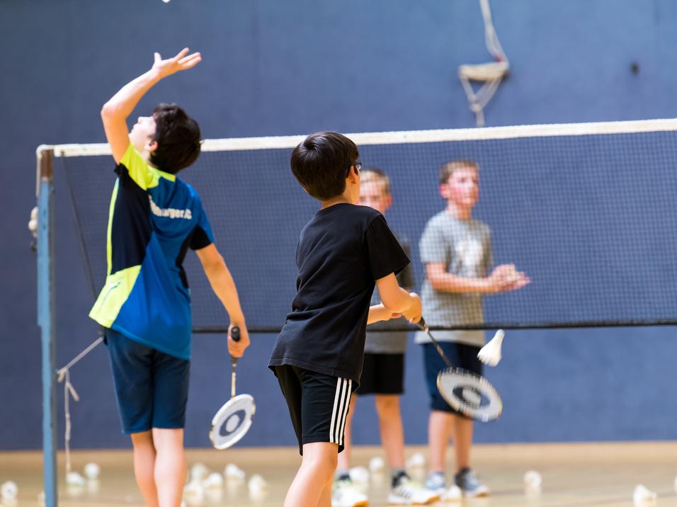 Gruppe von Kindern beim Badmintontraining in einer Sporthalle, mit Schlägern und Federbällen auf dem Boden.