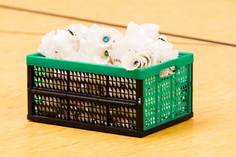 Ein grüner Korb mit vielen weißen Federbällen steht auf einem Holzfußboden in einer Sporthalle.