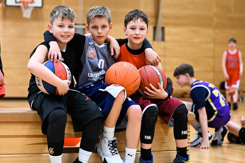 Drei Kinder posieren auf einer Bank in einer Sporthalle, jeder hält einen Basketball. Im Hintergrund weitere Spieler.
