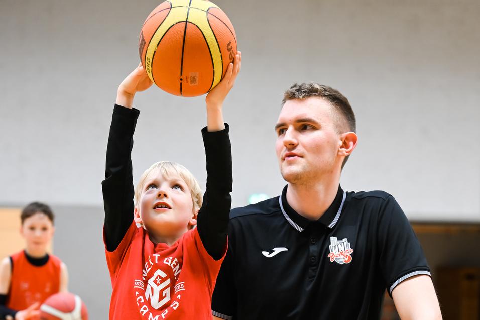 Ein junger Basketballspieler hebt den Ball über seinen Kopf, während ein Trainer neben ihm kniet und zuschaut.