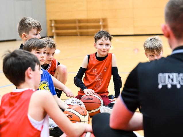 Gruppe von Jungen beim Basketballtraining, sitzend auf dem Boden mit Bällen, Trainer spricht zu ihnen.