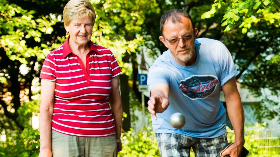Vier Personen stehen im Freien und beobachten, wie ein Mann eine Kugel auf einen Untergrund wirft.