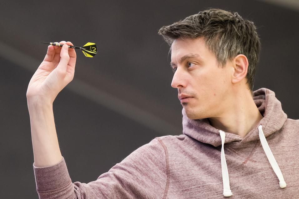 Ein Mann in einem grauen Hoodie zielt mit einem Dart auf eine Dartscheibe in einer Turnierumgebung.