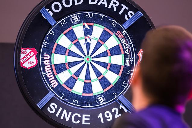 Ein Spieler wirft einen Pfeil auf ein beleuchtetes Dartboard mit dem Schriftzug „GOOD DARTS“ im Hintergrund.