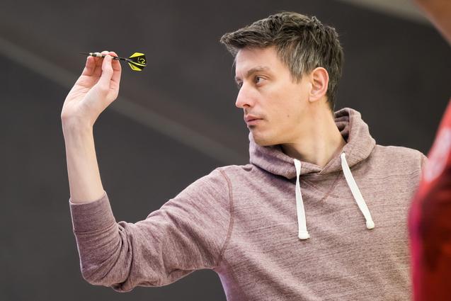 Ein Mann in einem grauen Hoodie zielt mit einem Dart auf eine Dartscheibe in einer Turnierumgebung.