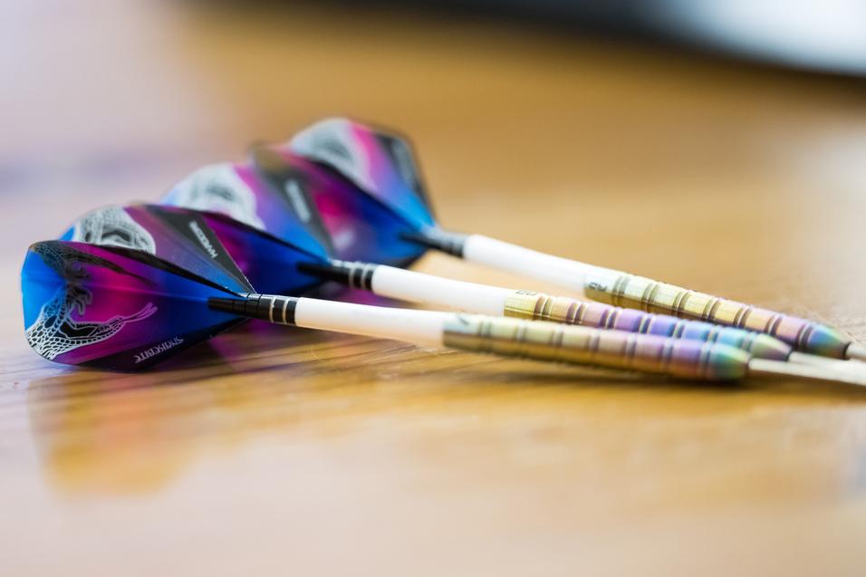 Drei Dartpfeile mit bunten Flights liegen auf einem Holztisch.