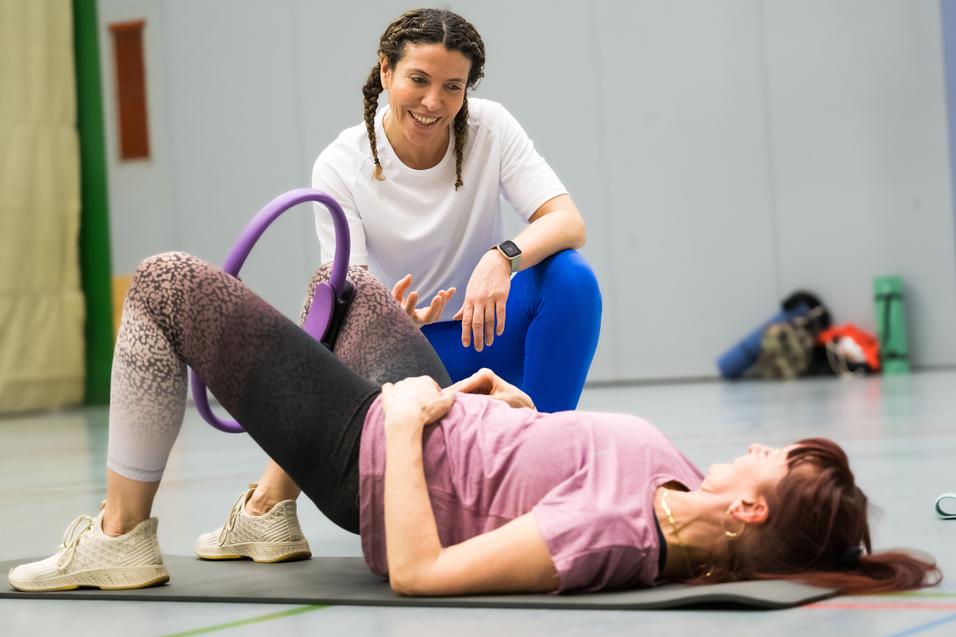 Zwei Frauen in Sportkleidung trainieren auf einer Matte, eine unterstützt die andere mit einem Fitnessgerät.