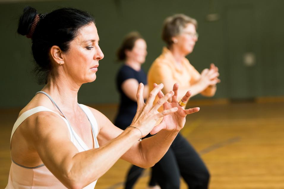 Drei Frauen führen eine Fitnessübung in einem Saal mit Holzfußboden und einer Sporttorwand im Hintergrund aus.