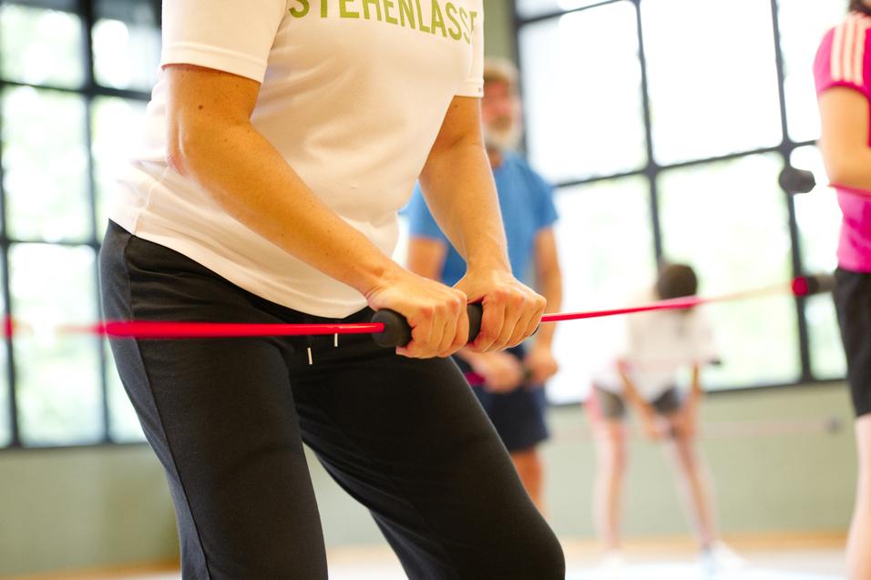 Nahaufnahme einer Person mit einem Widerstandsband im Fitnessstudio, umgeben von weiteren trainierenden Personen.