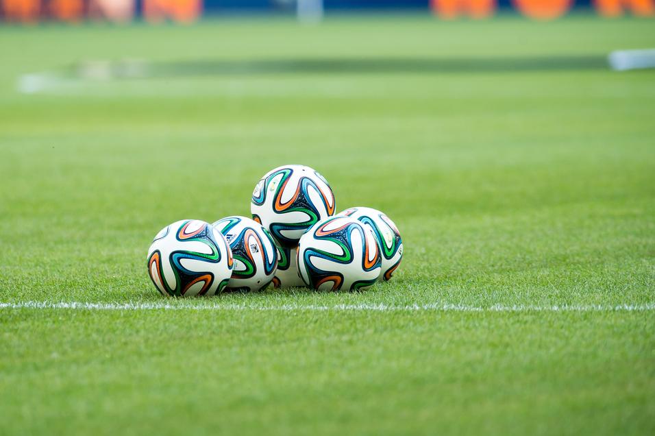 Fünf Fußballbälle liegen auf einem grünen Rasenfeld, sichtbar sind auch der Spielfeldrand und eine verschwommene Hintergrundszene.