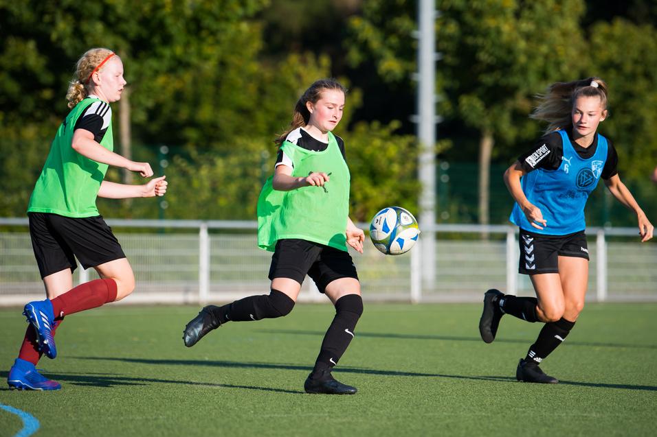 Drei Fußballspielerinnen in Trainingswesten schießen einen Ball auf einem Kunstrasenplatz unter blauem Himmel.