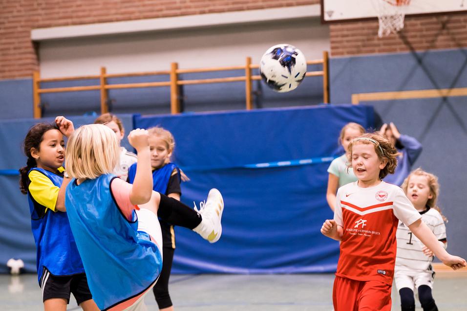 Mädchen spielen Fußball in einer Halle, ein Mädchen hebt den Fuß zum Schuss, während andere zuschauen.