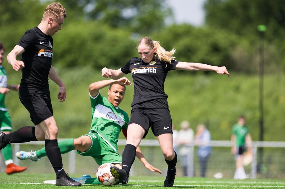 Drei Fußballspieler in Aktion auf einem Rasenplatz, eine Spielerin in Schwarz springt über einen am Boden liegenden Spieler.