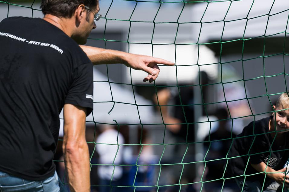 Ein Kind schaut durch ein Fußballtor, während ein Erwachsener auf es zeigt und ein Ball in der Hand hält.