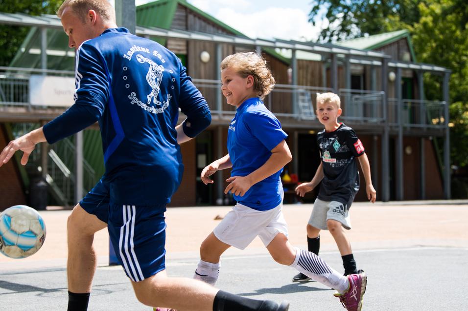 Zwei Kinder in Sportkleidung und ein Trainer spielen Fußball auf einem Platz mit Gebäuden im Hintergrund.