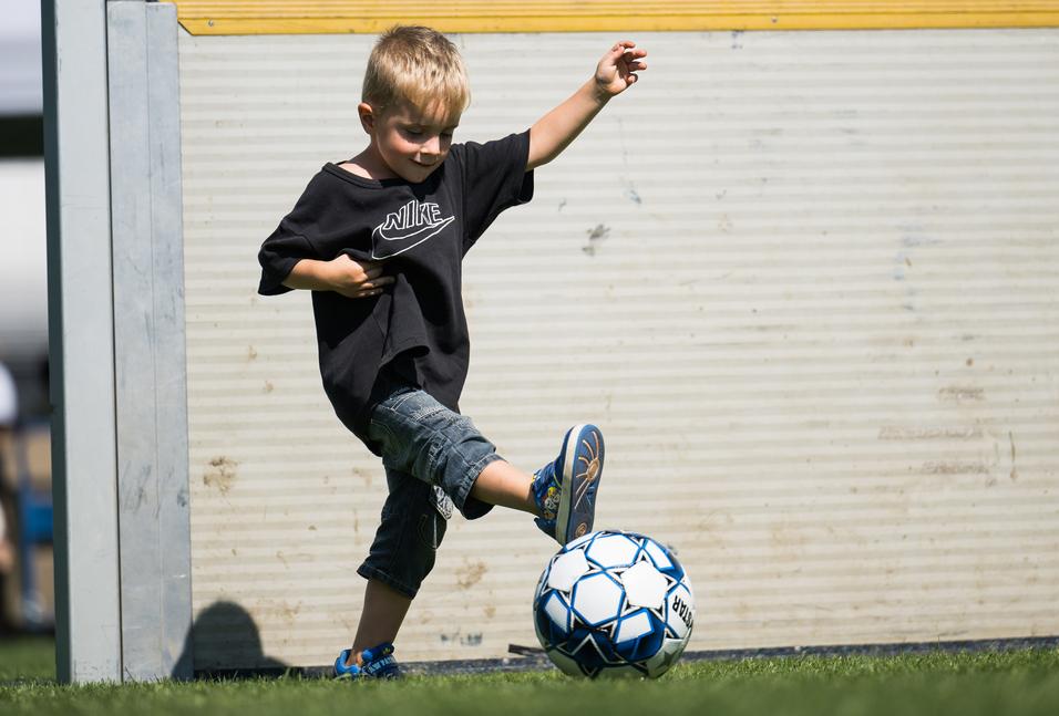 Ein kleiner Junge in einem schwarzen T-Shirt tritt auf einen Fußball vor einem Tor auf einem Rasenplatz.