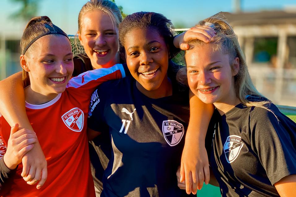 Vier junge Fußballspielerinnen in Teamtrikots posieren lachend gemeinsam auf dem Spielfeld.