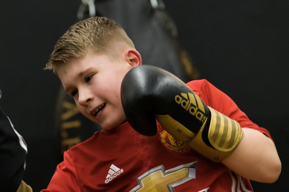 Junge in rotem Trikot mit Boxhandschuhen trainiert mit einem Trainer an einem Boxsack.