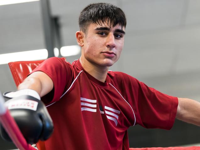 Jugendlicher Boxer in rotem Shirt lehnt an einem Boxring, trägt Handschuhe und schaut direkt in die Kamera.