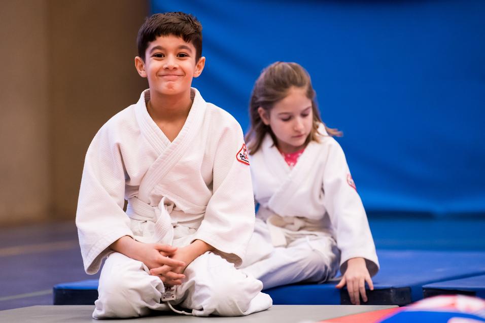 Zwei Kinder in weißen Judoanzügen sitzen auf einer Matte in einem Judoraum, der Hintergrund ist blau.