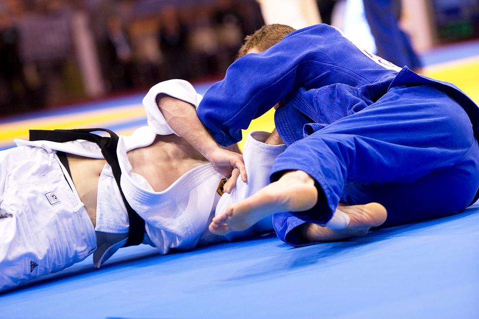 Zwei Judoka kämpfen auf einer blauen Matte, einer in weißem und der andere in blauem Gi, während Zuschauer im Hintergrund zuschauen.