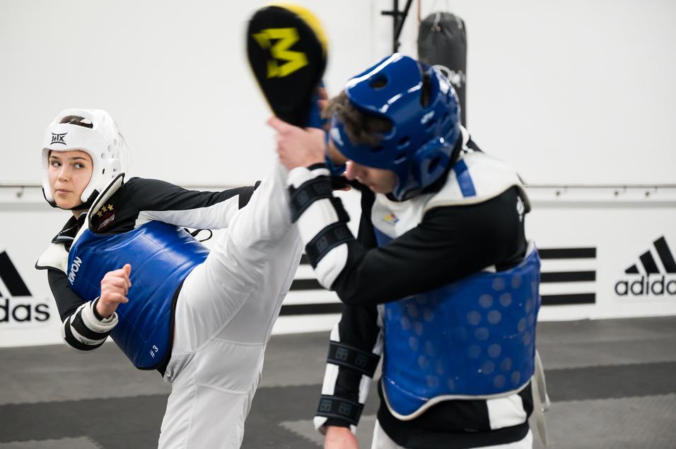 Zwei Kampfkämpfer im Taekwondo-Training; eine Person führt einen hohen Kick aus, beide tragen Schutzausrüstung.