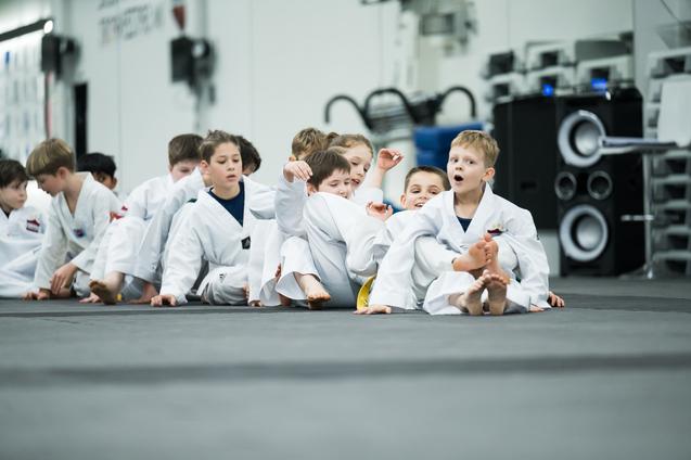 Gruppe von Kindern in weißen Taekwondo-Anzügen sitzt auf einer Matte und interagiert freundlich miteinander.