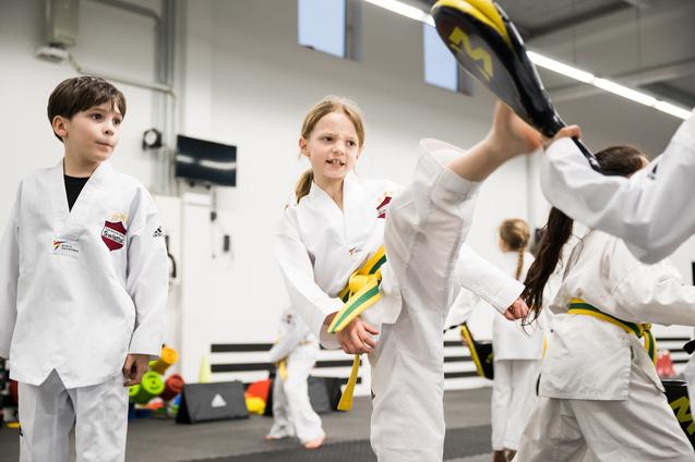 Kinder in weißen Taekwondo-Anzügen trainieren, während ein Mädchen mit gelbem Gürtel einen hohen Kick ausführt.