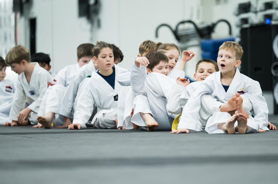 Gruppe von Kindern in weißen Taekwondo-Anzügen sitzt auf einer Matte und interagiert freundlich miteinander.