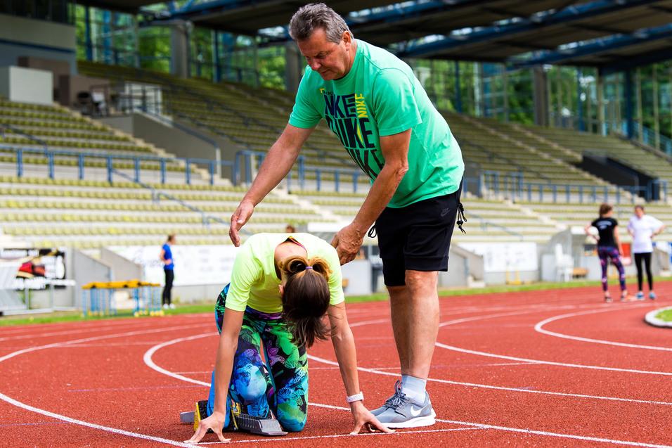 Trainer hilft Sportlerin beim Startblock auf einer Laufbahn in einem Stadion. Im Hintergrund stehen weitere Athleten.