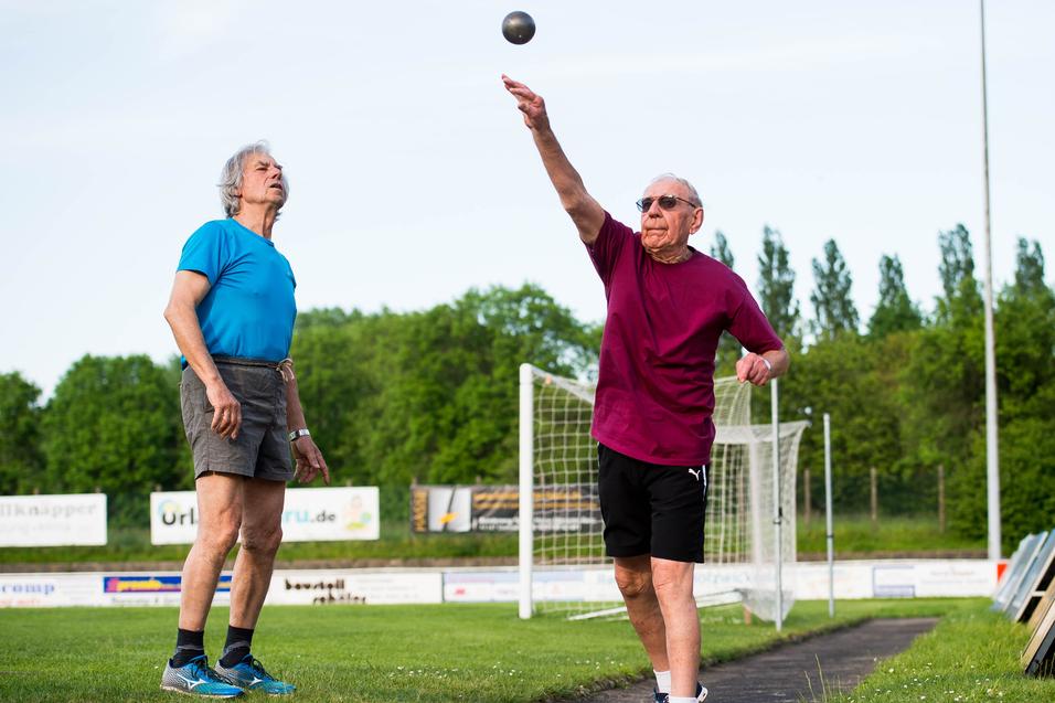 Zwei ältere Männer üben das Kugelstoßen auf einem Sportplatz, einer wirft die Kugel, der andere beobachtet.