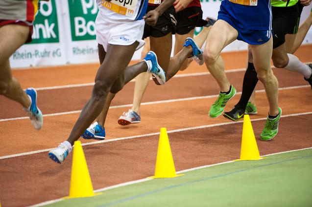 Beine von mehreren Läufern in Wettkampfkleidung, die auf einer Laufbahn um gelbe Kegel sprinten.