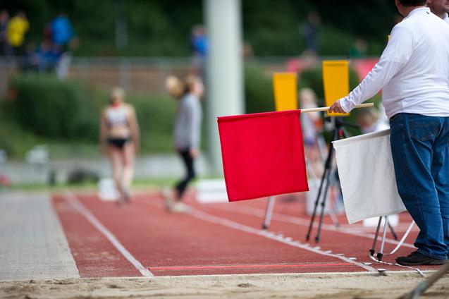 Ein Schiedsrichter hält eine rote Flagge neben einer Laufbahn, während Athleten im Hintergrund vorbeigehen.