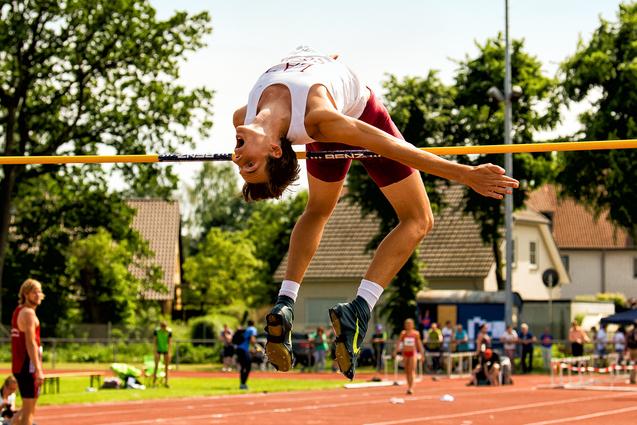 Weitspringender Athlet in Aktion, der über eine hohe Latte springt, mit Zuschauern im Hintergrund auf einem Sportplatz.