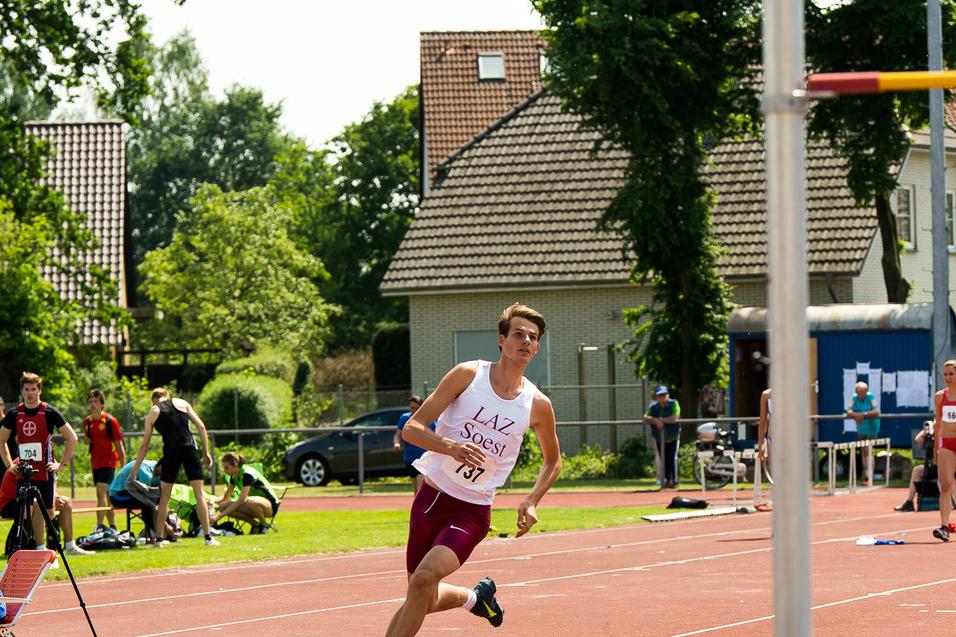 Athlet in Sportkleidung sprintet auf einer Leichtathletikbahn, im Hintergrund Zuschauer und eine Hochsprung-Anlage.