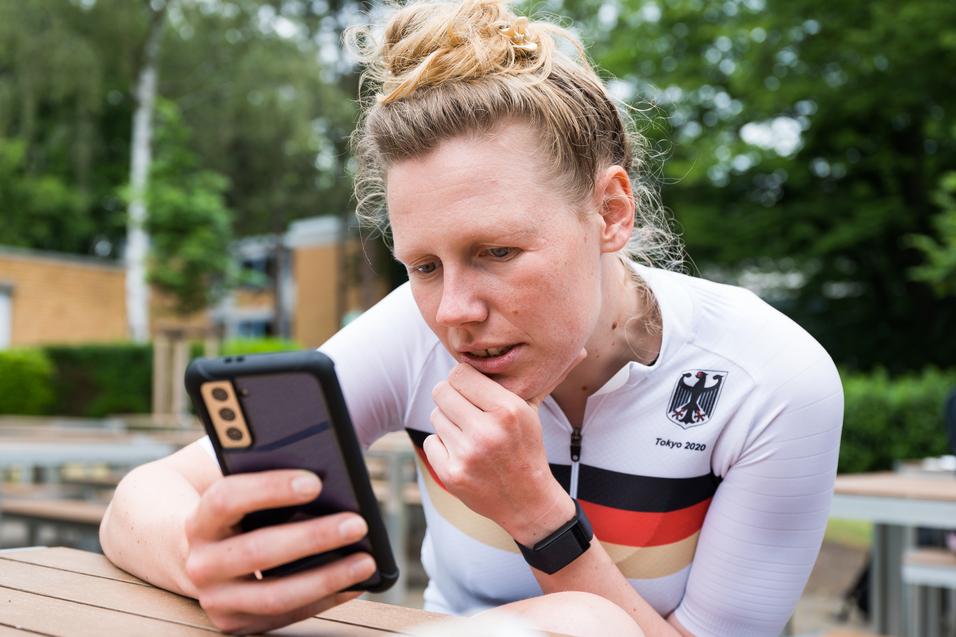 Athletin im weißen Trikot mit schwarzem und rotem Streifen schaut konzentriert auf ihr Smartphone in einer Außenanlage.