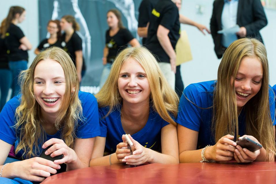 Drei lachende Jugendliche in blauen T-Shirts liegen auf einem Tisch und schauen auf ihre Handys. Hintergrund mit weiteren Personen.