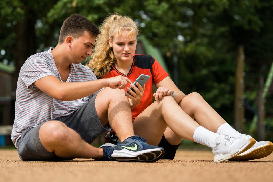 Zwei Jugendliche sitzen auf dem Boden und betrachten gemeinsam ein Smartphone. Ein Mädchen trägt ein rotes sportliches Oberteil.