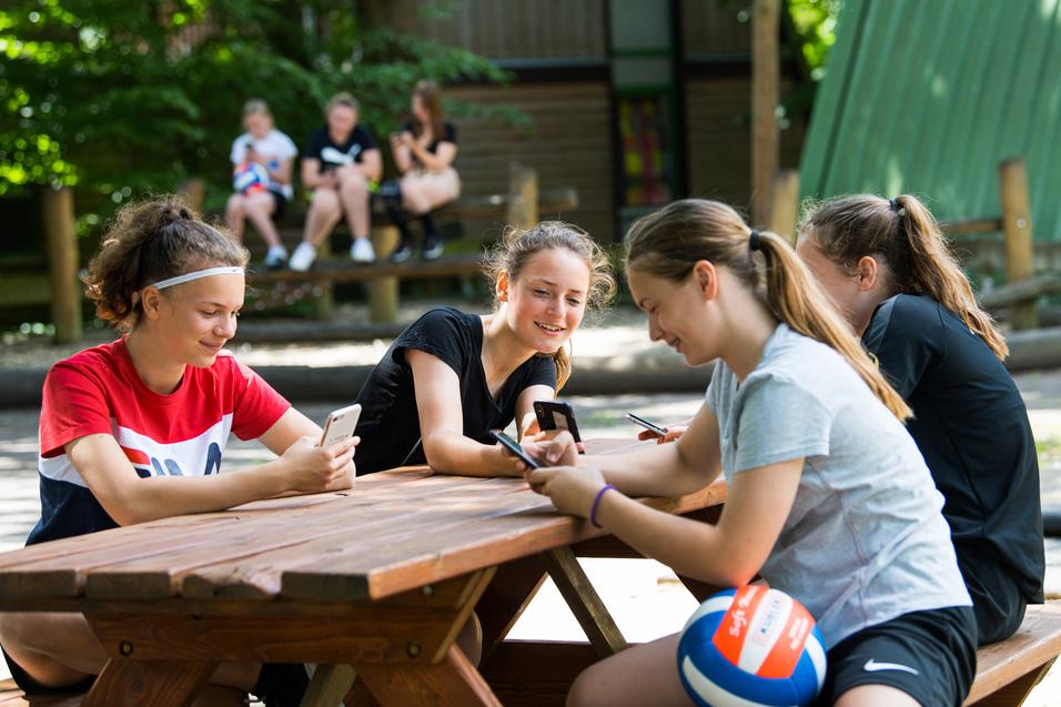 Vier Mädchen sitzen an einem Holztisch und nutzen ihre Smartphones, während im Hintergrund andere Kinder spielen.