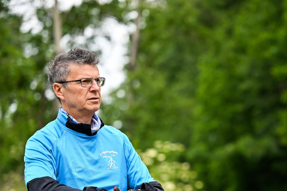 Mann in blauer Sportjacke hält Nordic Walking-Stöcke in einer grünen, bewaldeten Umgebung.