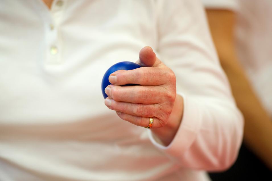 Nahaufnahme einer Hand, die einen blauen Ball hält, während die Person in einem weißen Oberteil sitzt.