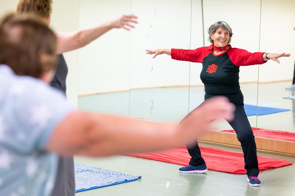 Eine ältere Frau in Sportkleidung führt eine Dehnübung in einem Fitnessraum mit Spiegeln aus.