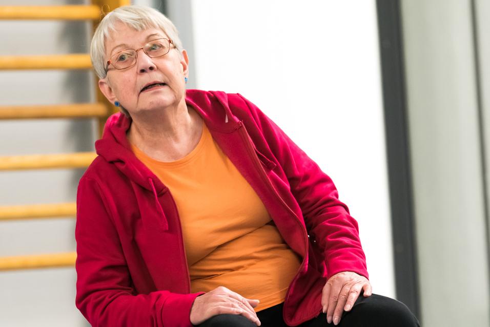 Ältere Frau in roter Jacke sitzt auf einem blauen Gymnastikball in einer Sporthalle.