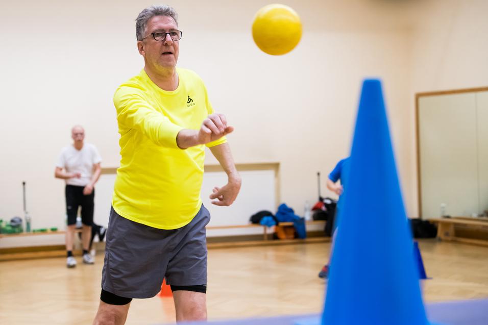Ein Mann in gelber Sportkleidung wirft einen Ball auf einen blauen Kegel in einem Übungsraum. Hintergrund unscharf.