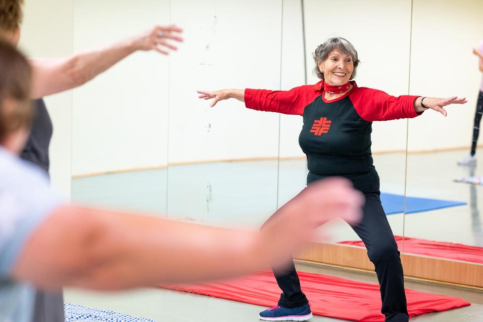 Eine ältere Frau in Sportkleidung führt eine Dehnübung in einem Fitnessraum mit Spiegeln aus.