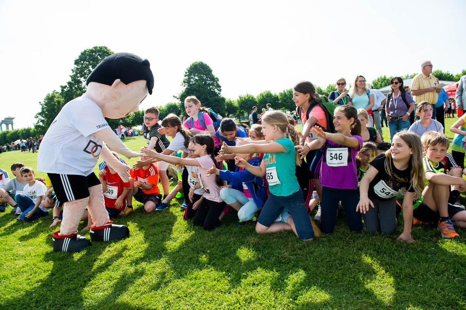 Kinder in Sportbekleidung strecken die Hände nach einem großen Maskottchen in einem grünen Park aus.