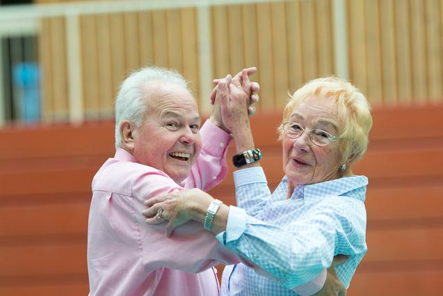 Ein älteres Paar tanzt lächelnd in einer Turnhalle, Kleidung in sanften Farben. Holzpaneele im Hintergrund.