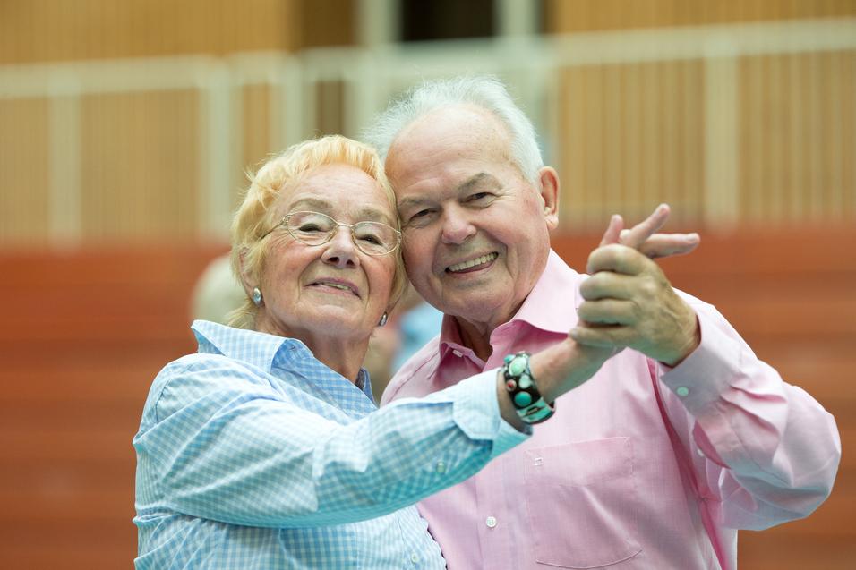 Ein älteres Paar umarmt sich lächelnd und tanzt in einer Sporthalle. Ihre Kleidung ist hell und farbenfroh.