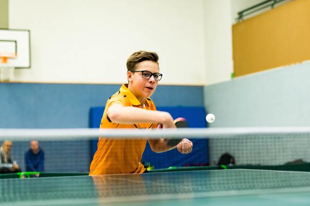 Junger Tischtennisspieler in orangefarbenem Shirt schlägt den Ball auf einem Tischtennistisch.